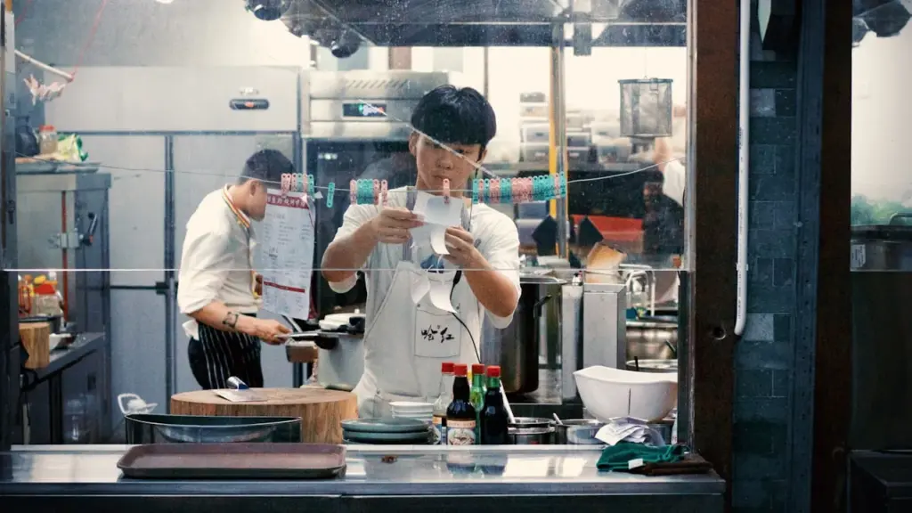 A professional kitchen briefing scene, head chef speaking to attentive staff around a stainless steel prep counter, warm.