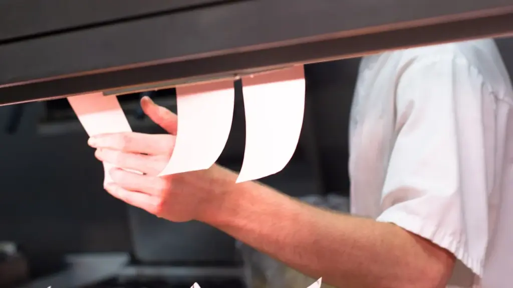 A professional kitchen pass at full service, stainless steel counter with plated dishes, a chef in whites coordinating.
