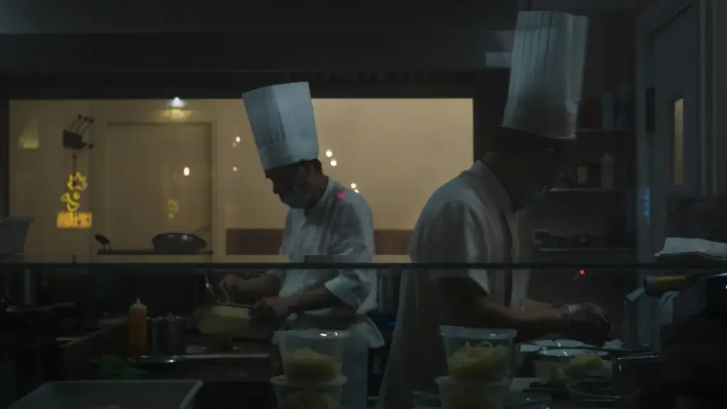 A professional kitchen interview scene, two people across a stainless steel counter, warm overhead lighting, serious and.