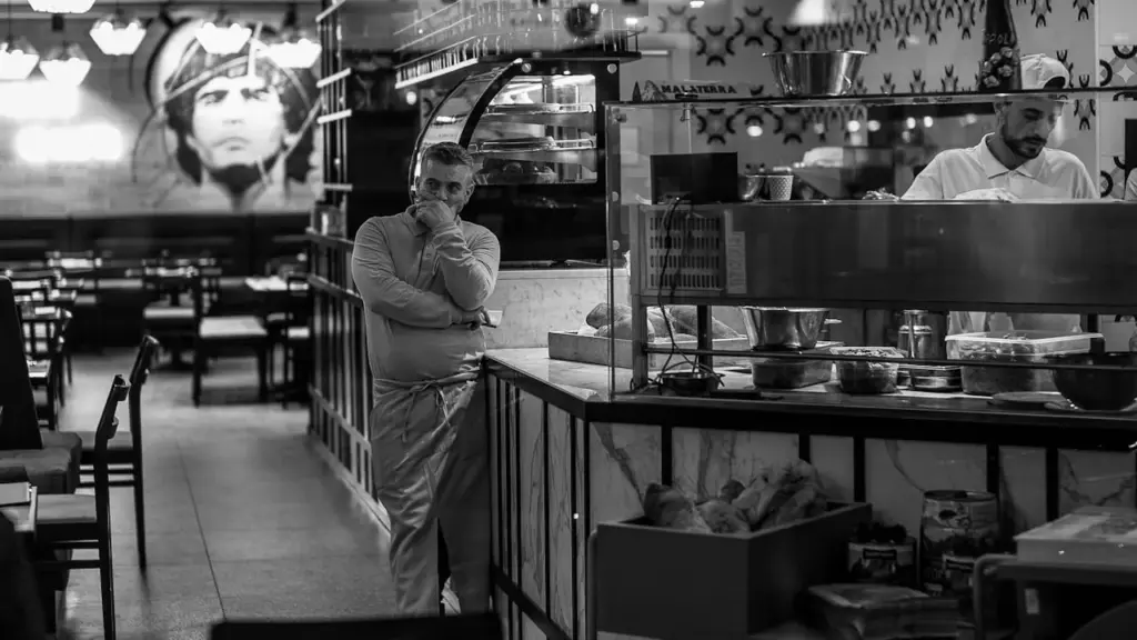 A professional restaurant kitchen pass at peak service, warm amber lighting, a chef and front of house staff making brief.