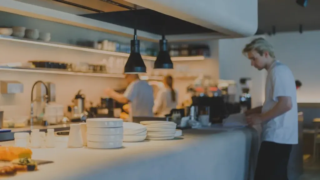 Two chefs in a professional kitchen, one confident and focused, one technically precise but tense, warm overhead lighting