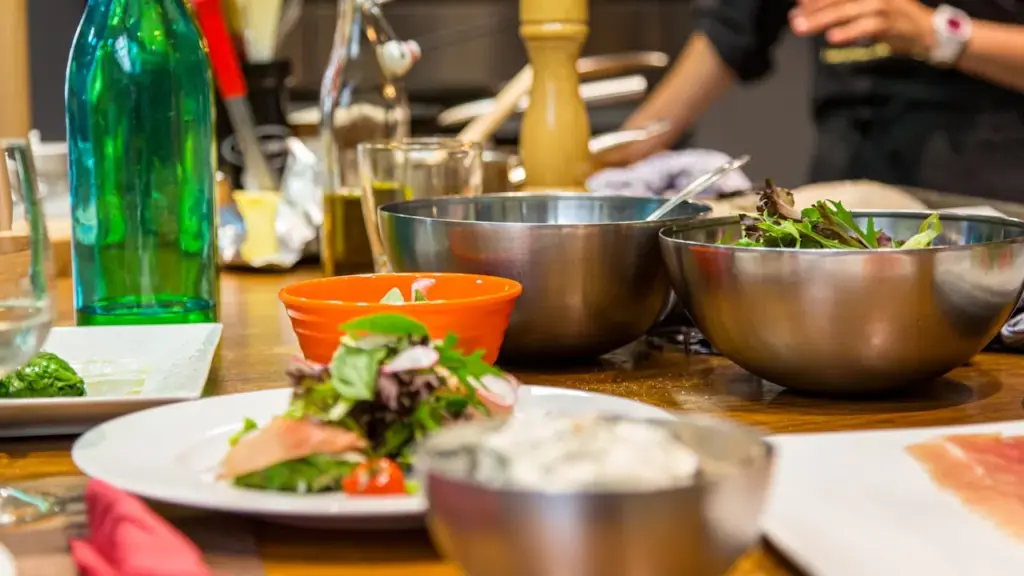 A long stainless steel kitchen prep table with mismatched bowls of hearty food, steam rising, chefs seated together.