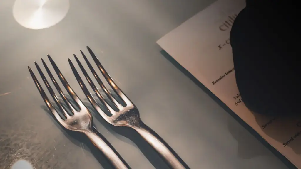 A restaurant kitchen pass at night, two plated dishes side by side, dramatic overhead light, one dish richly garnished, one.