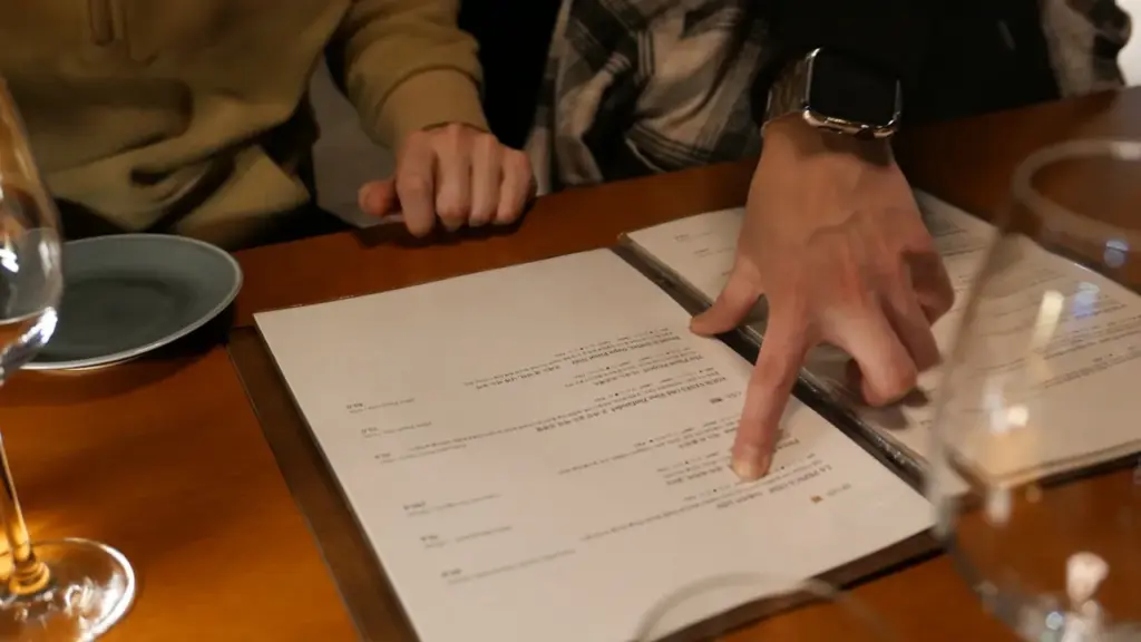 Overhead shot of a chef reviewing handwritten recipe cost sheets beside fresh ingredients on a worn kitchen prep table, warm.