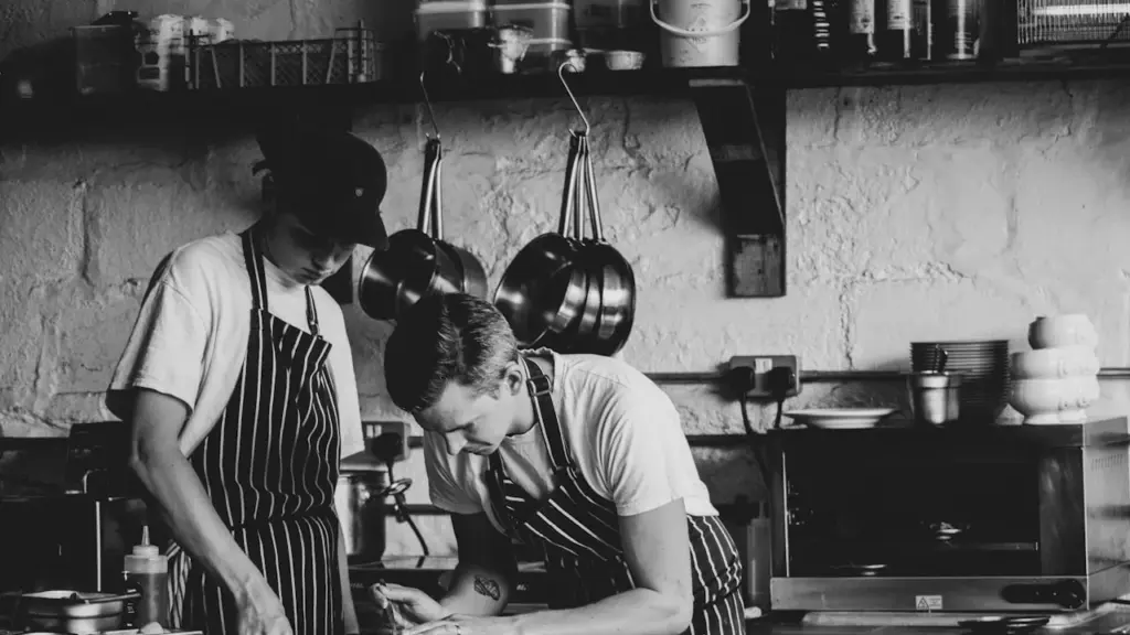 Two chefs in a professional kitchen, one elder guiding a younger cook with calm authority, warm ambient light