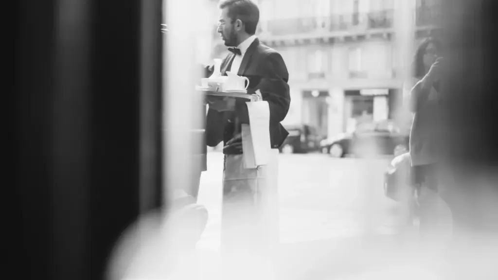 A composed head waiter in formal attire surveying a busy restaurant dining room at dusk, warm candlelight